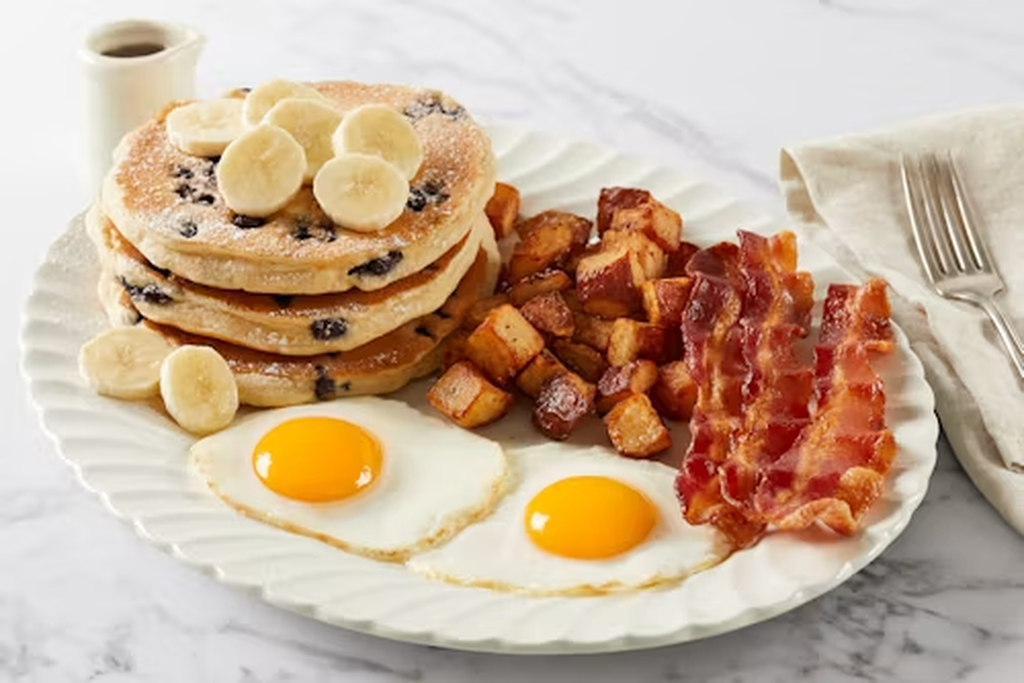 Banana Berry Farmer breakfast with blueberry pancakes topped with fresh banana slices, two sunny-side-up eggs, crispy bacon and seasoned breakfast potatoes served with syrup.